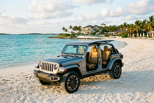 Silver open-top Jeep Wrangler parked on a white sand beach with turquoise water.