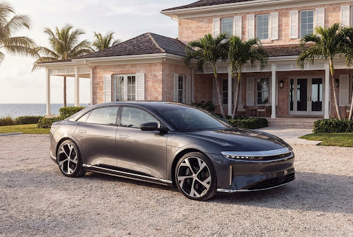 Sleek grey Lucid electric sedan parked on a gravel driveway in front of a luxury island villa.